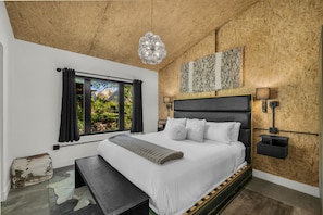 A bedroom with a large bed, a bench, a window with curtains, and a wall-mounted lamp.