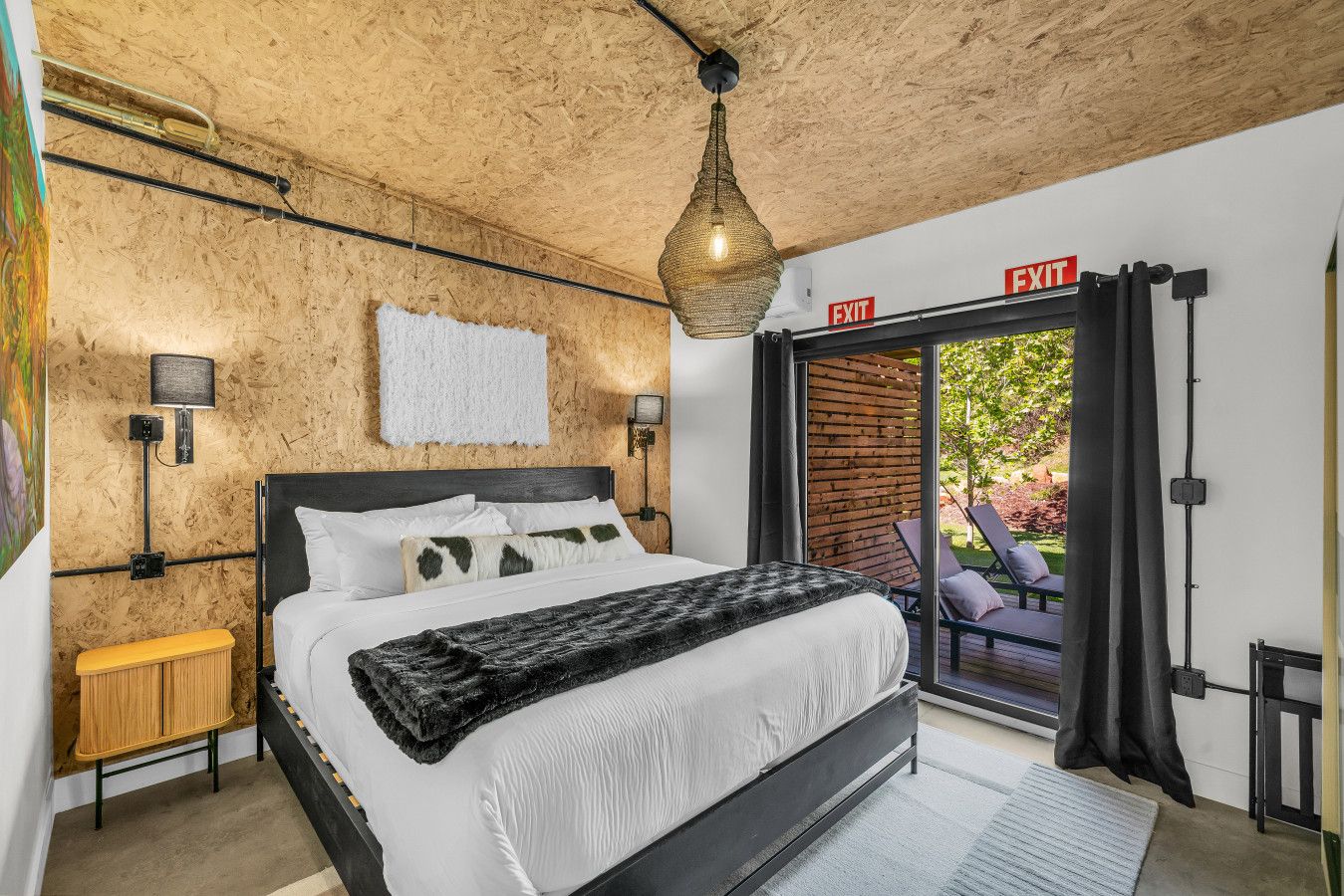 Ein Schlafzimmer mit einem Bett, einem Nachttisch, einer Lampe, einem Fluchtwegschild und Blick ins Freie.