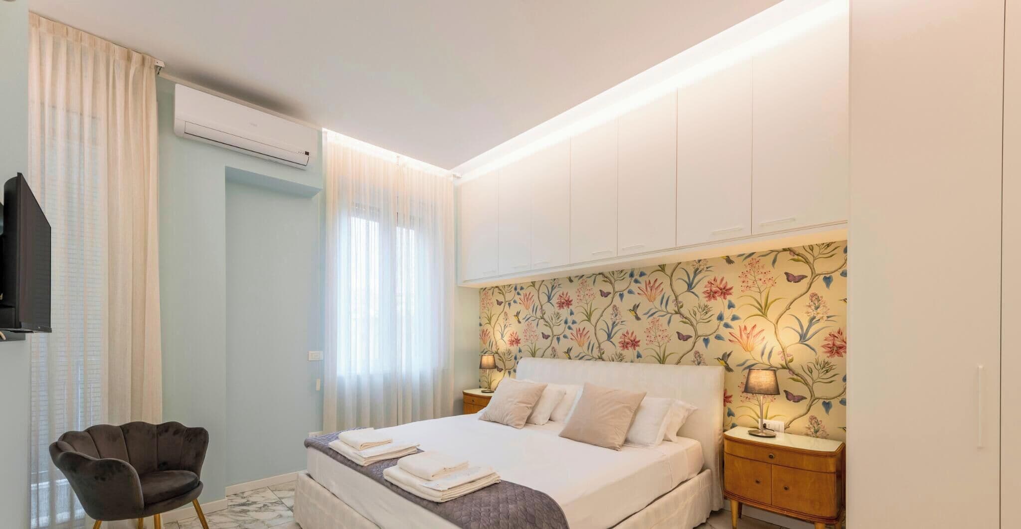 A bedroom with a bed, bedside tables, a chair, an air conditioning unit, and a television.