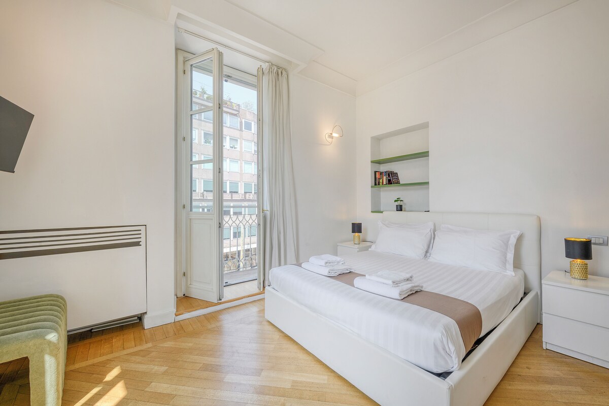 A hotel room with a bed, a bedside table, a nightstand, a chair, a window with curtains, and a view of buildings outside.