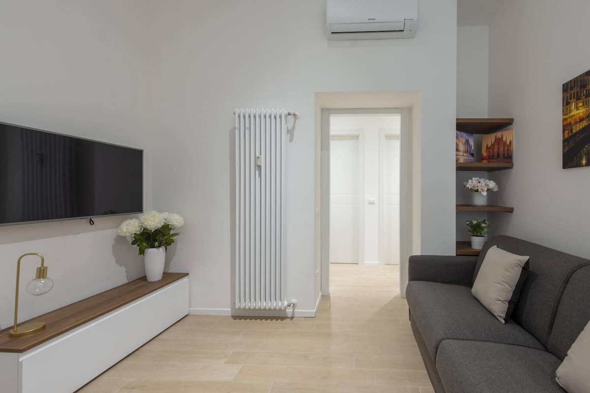 A modern living room with a flat-screen TV, a radiator, a sofa, and a console table with a lamp and flowers.