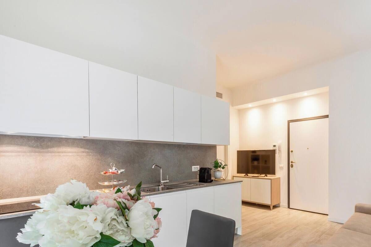 A modern kitchen with white cabinets, a sink, and a countertop with a coffee maker and a plant. There is a dining area with a table and chairs, and a sofa in the living room.