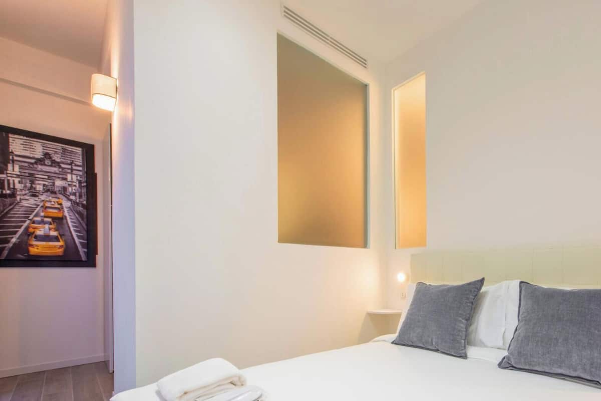 A hotel room with a bed, pillows, a nightstand, a wall-mounted light, and a framed picture of a cityscape.