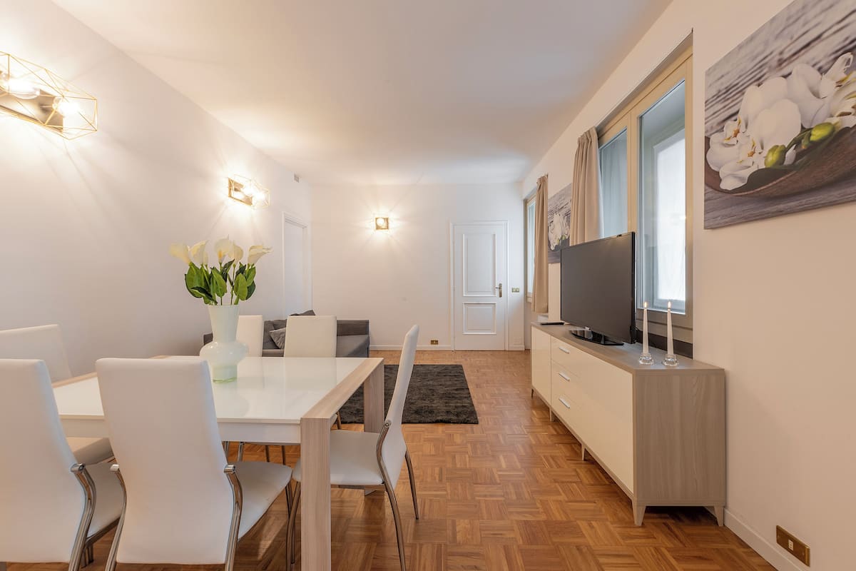 A modern kitchen with a built-in oven, a dining area with a white table and chairs, and a TV on a wooden cabinet.
