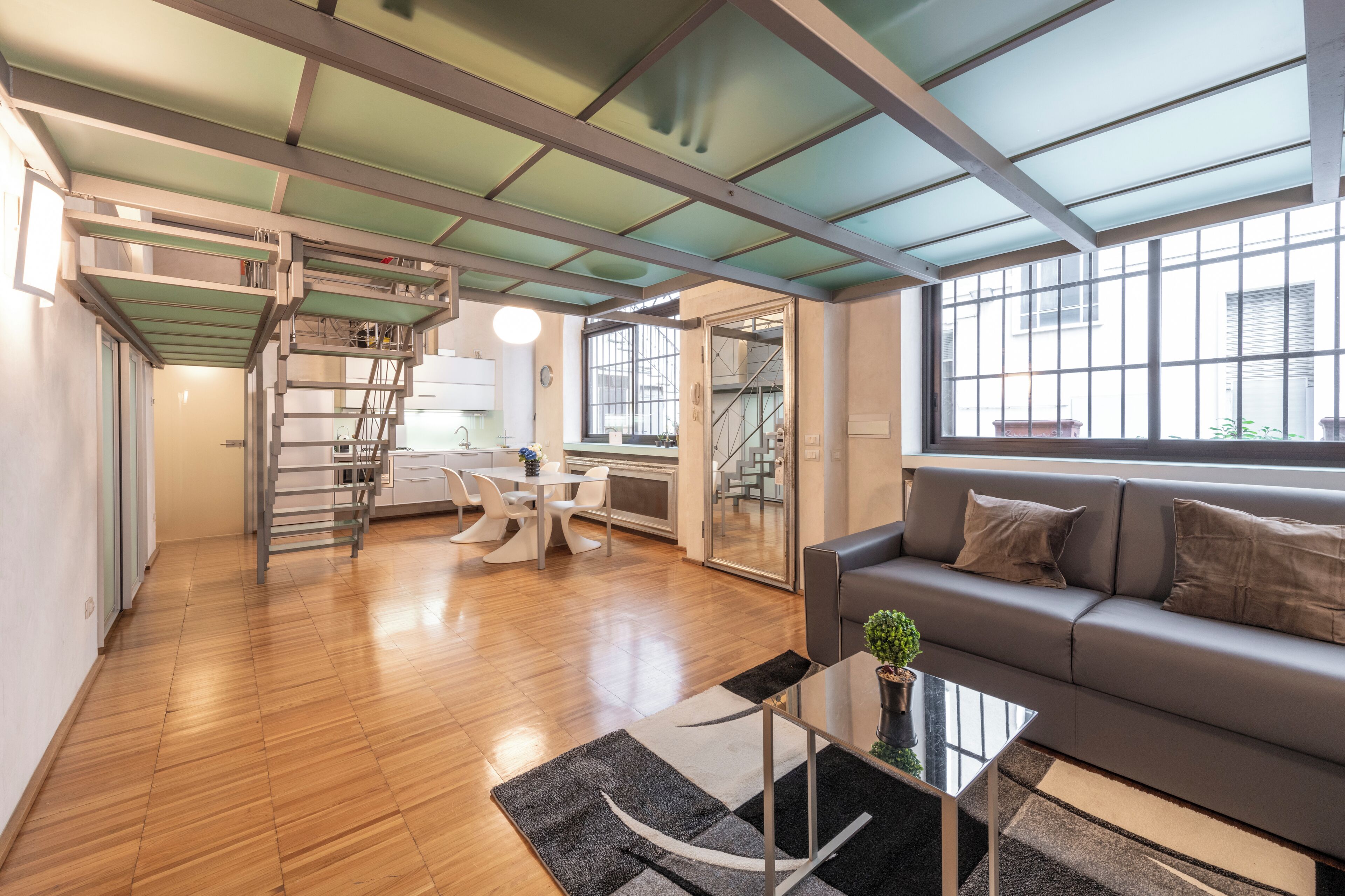 A modern living room with a wooden floor, a grey sofa, a glass coffee table, and a staircase leading to an upper level.