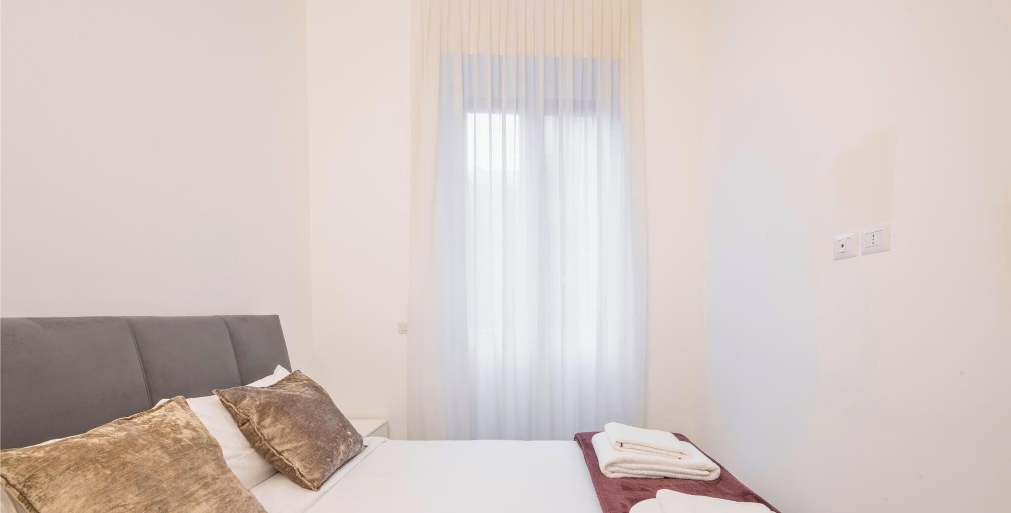 A hotel room with a bed, a nightstand, a window with sheer curtains, and a wall socket.