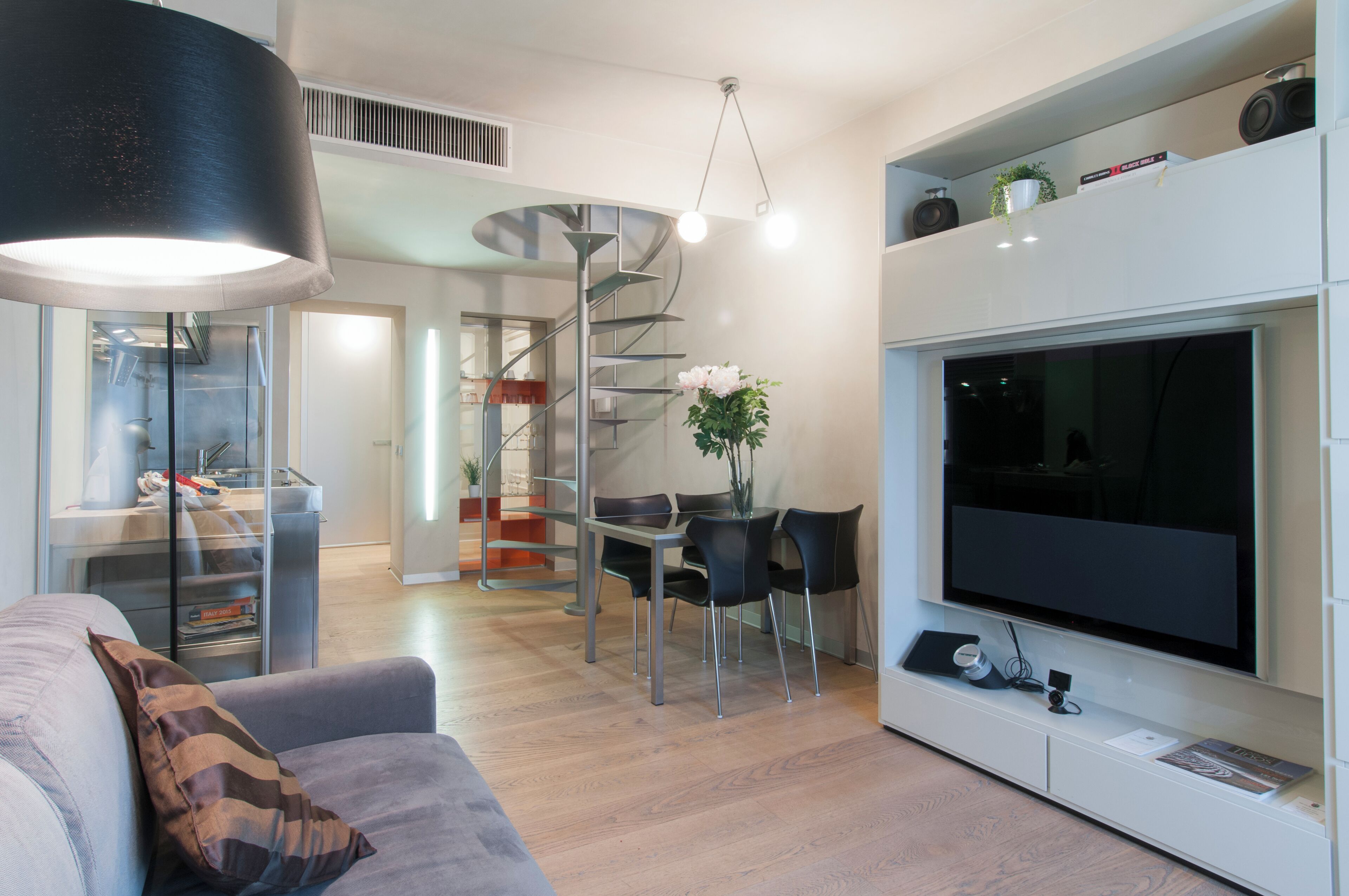 A modern living room with a flat-screen TV, a dining area with a spiral staircase, and a large black lamp.