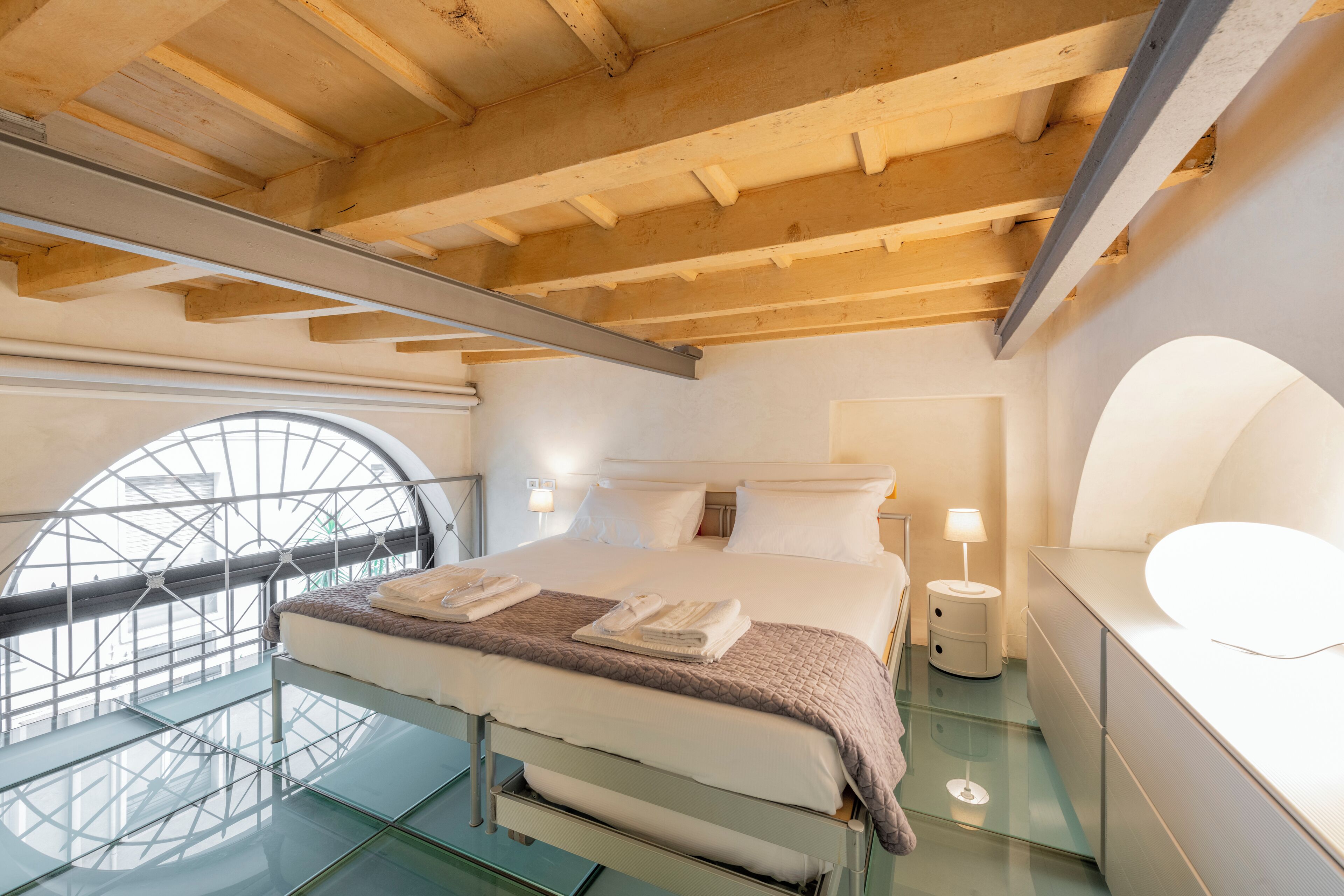 A modern hotel room with a glass floor, a large bed with white linens, a wooden ceiling, and a circular window.