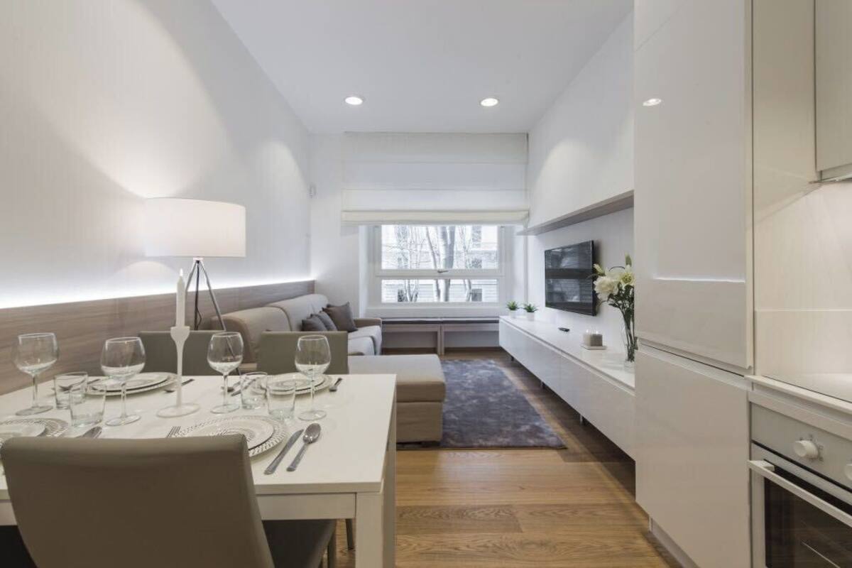 A modern dining area with a table set for four, a sofa, a TV mounted on the wall, and a window with a view of trees.