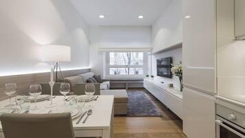 A modern dining area with a table set for four, a sofa, a TV mounted on the wall, and a window with a view of trees.