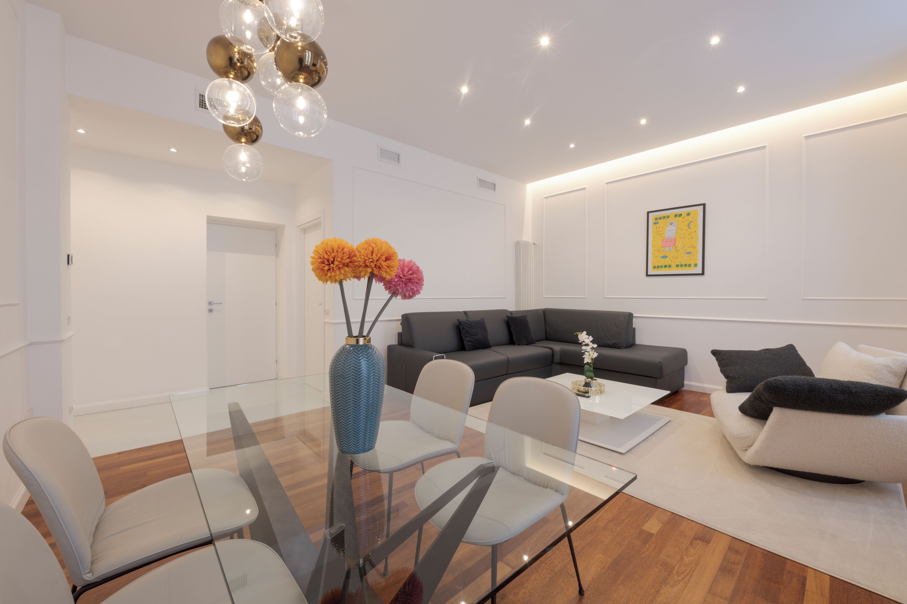 A modern living room with a glass dining table, grey sofas, and a blue vase with flowers.