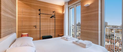 A modern hotel room with a large bed, a wooden wall panel, a view of the city, and a hanging coat rack.