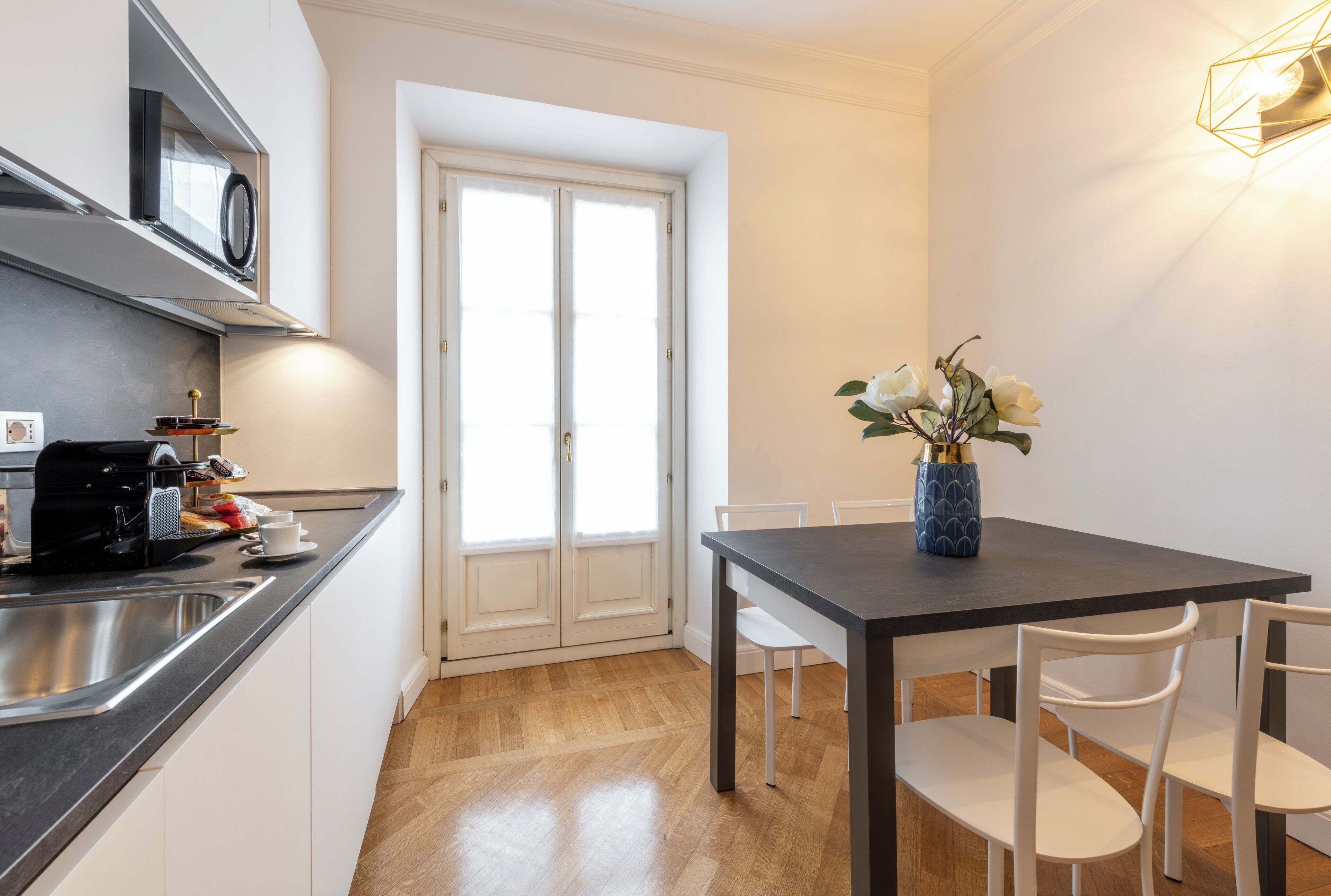 Une cuisine moderne équipée d’un four à micro-ondes encastré, d’une machine à café et d’un évier. On y trouve une table à manger avec des chaises blanches et un vase rempli de fleurs.