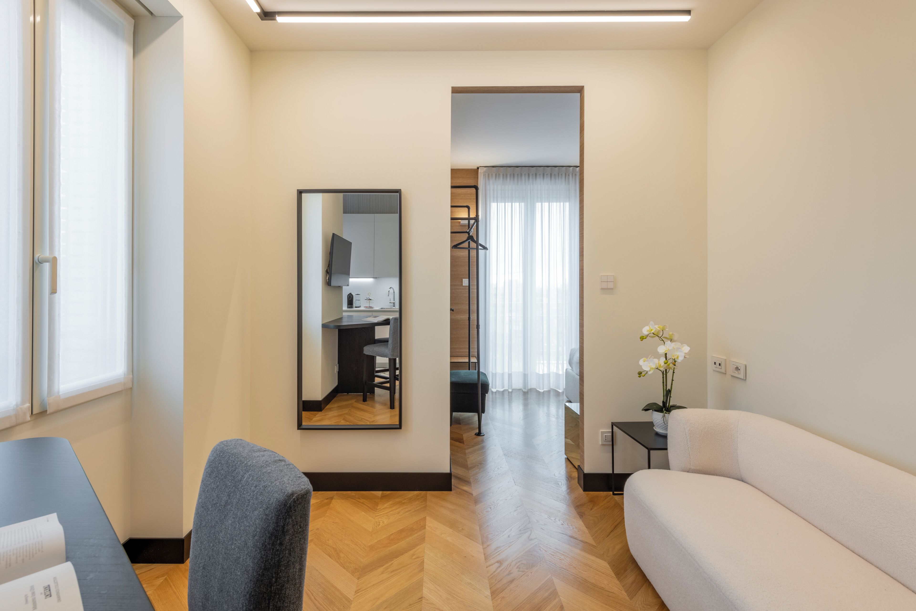 A modern living room with a sofa, a chair, a small table, and a mirror. There is a window with blinds, a door, and a hallway leading to a kitchen area.