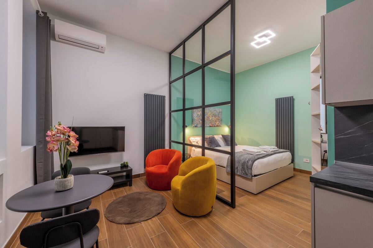 A modern room with a bed, a TV, a dining area with chairs, and a red and yellow chair.