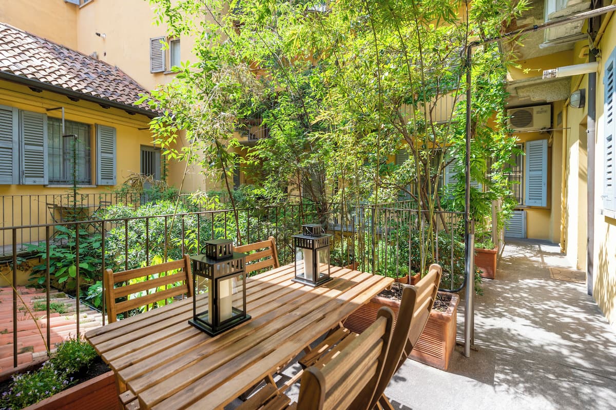 A wooden table with benches and lanterns in a courtyard surrounded by buildings with balconies and railings.