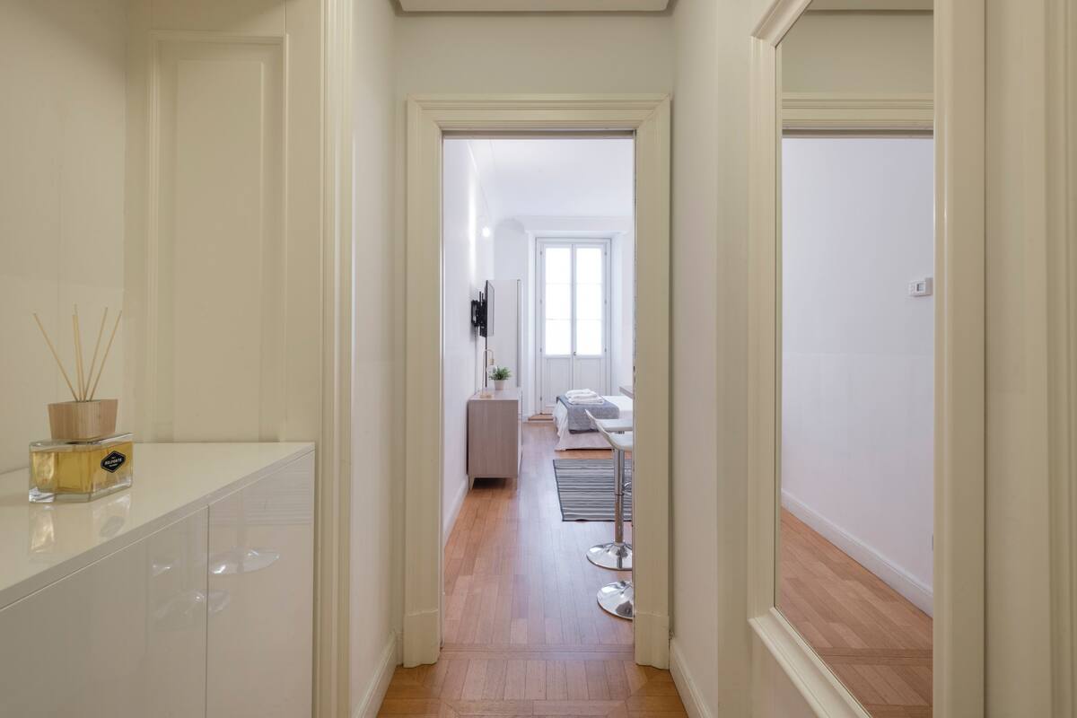 A hallway with a wooden floor leading to a bedroom with a bed, bedside table, and a window.