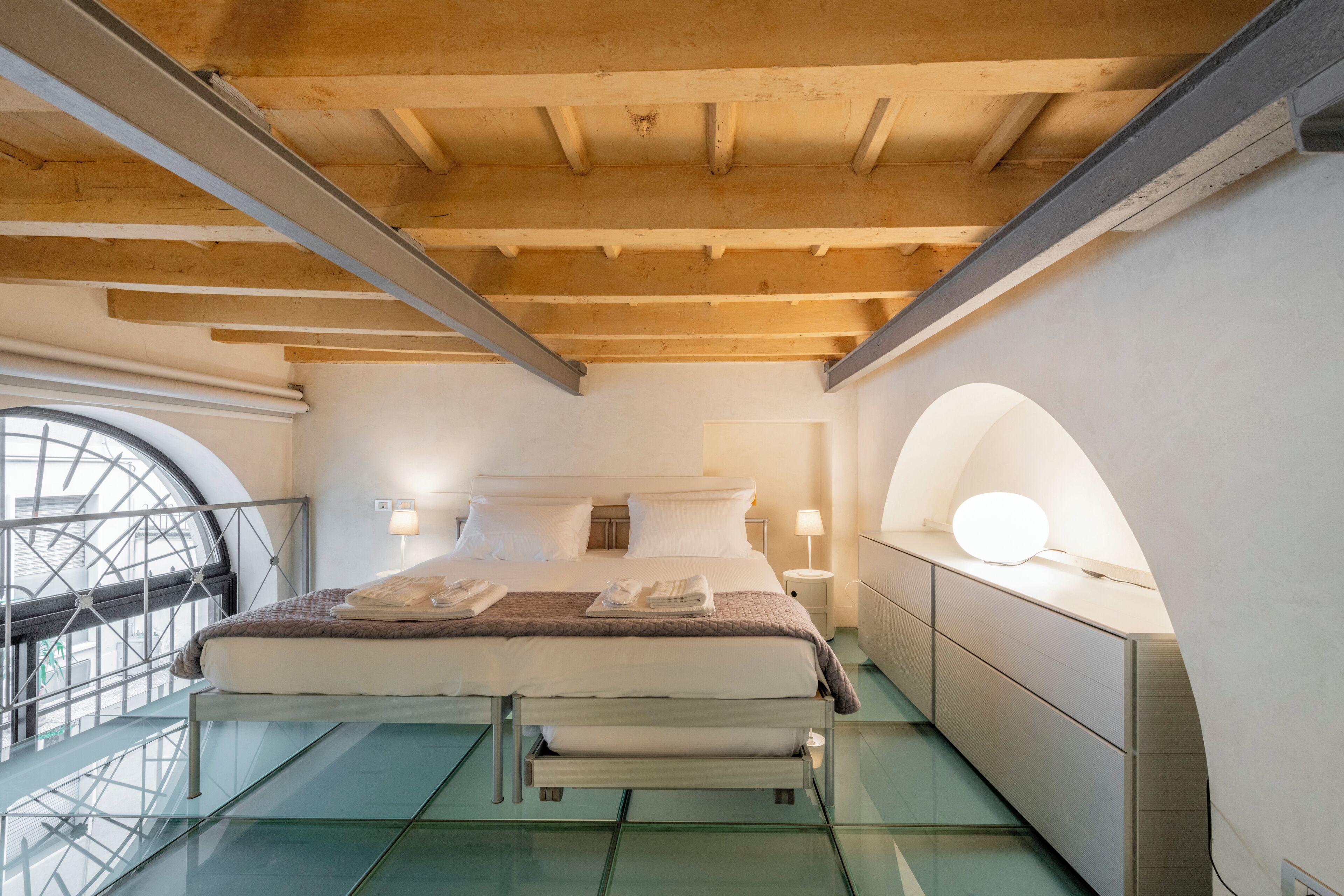 A modern bedroom with a glass floor, a bed with white linens, a round mirror, and a nightstand with a lamp.