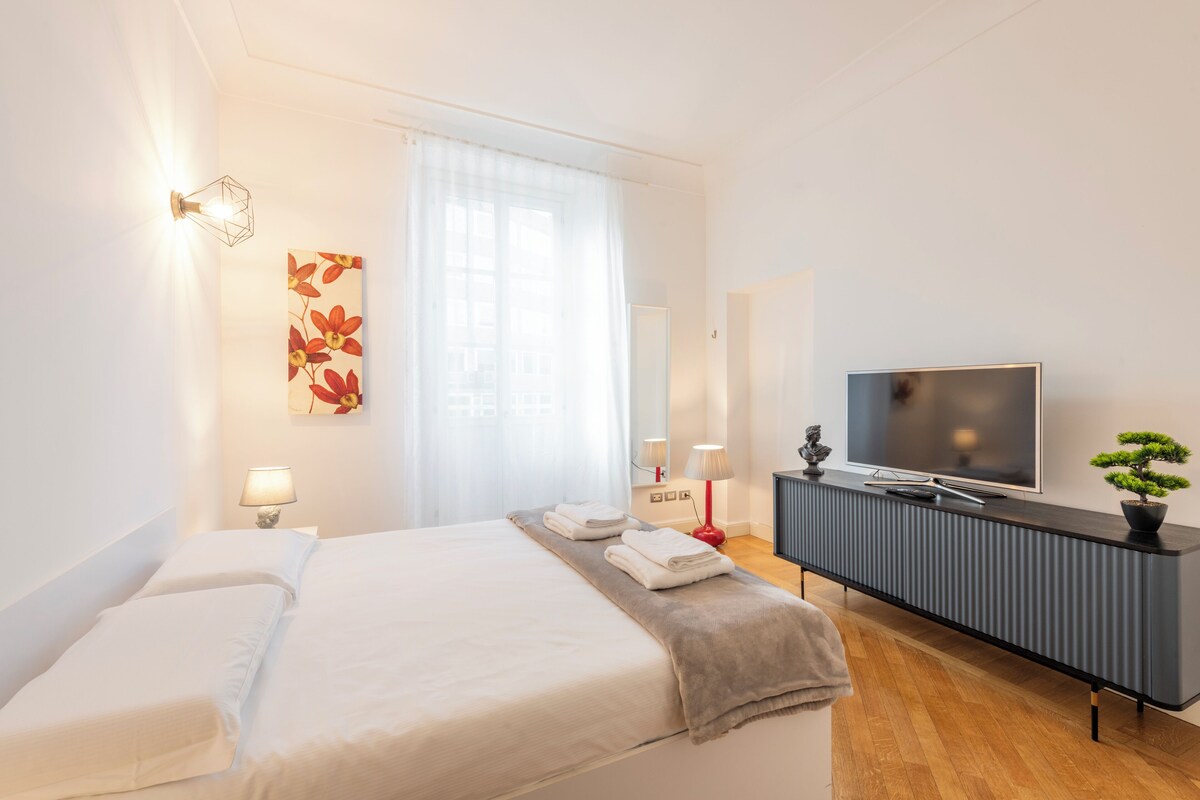 A modern hotel room with a large bed, a flat-screen TV, a bedside table with lamps, and a decorative wall piece.