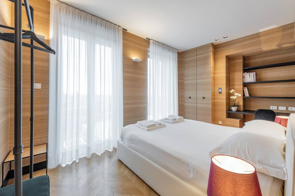 A modern hotel room with a large bed, a bedside table with a lamp, and a built-in bookshelf.