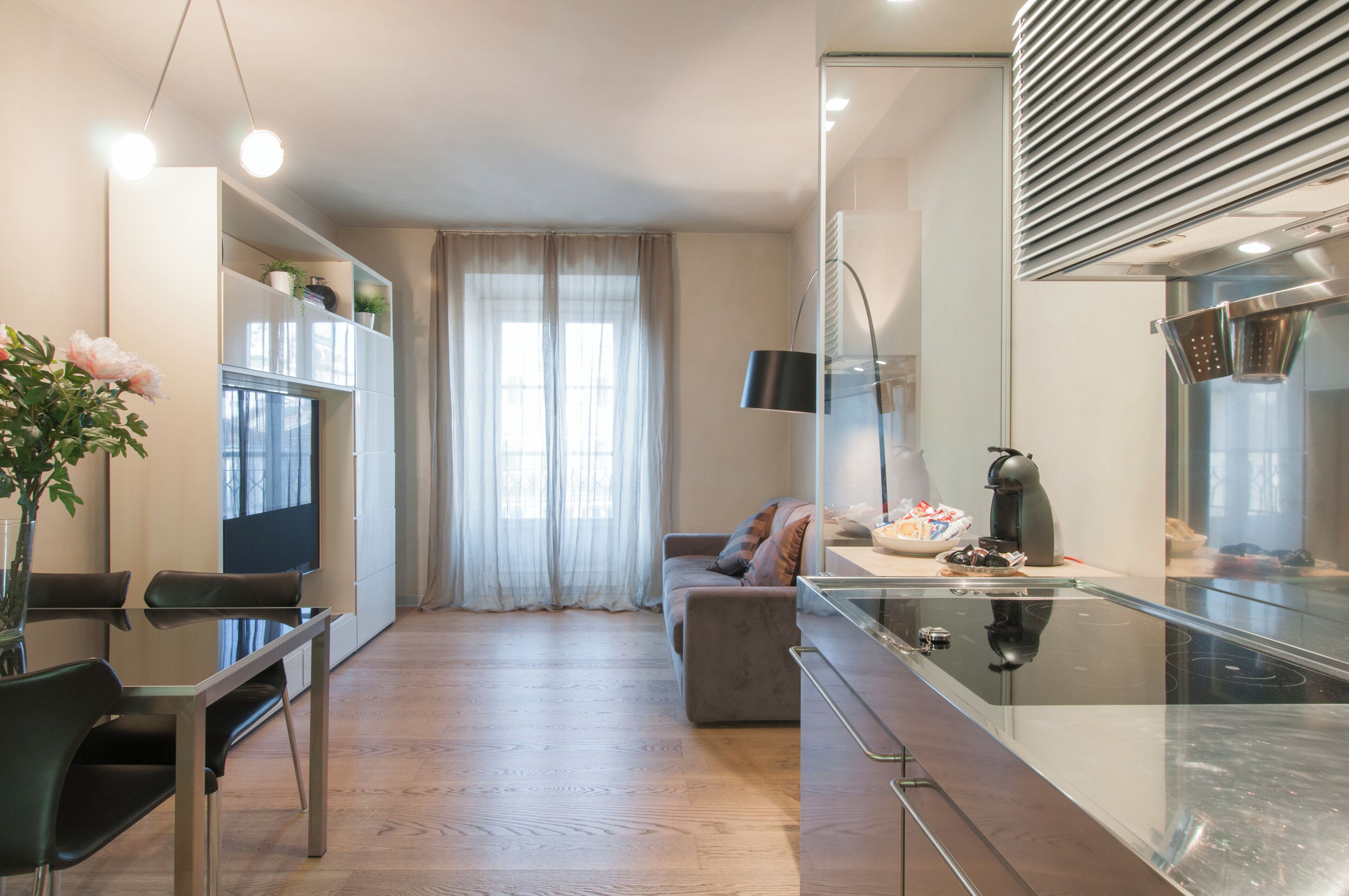 A modern kitchen with a built-in stove, a dining area with a glass table and chairs, and a living area with a sofa and a TV.