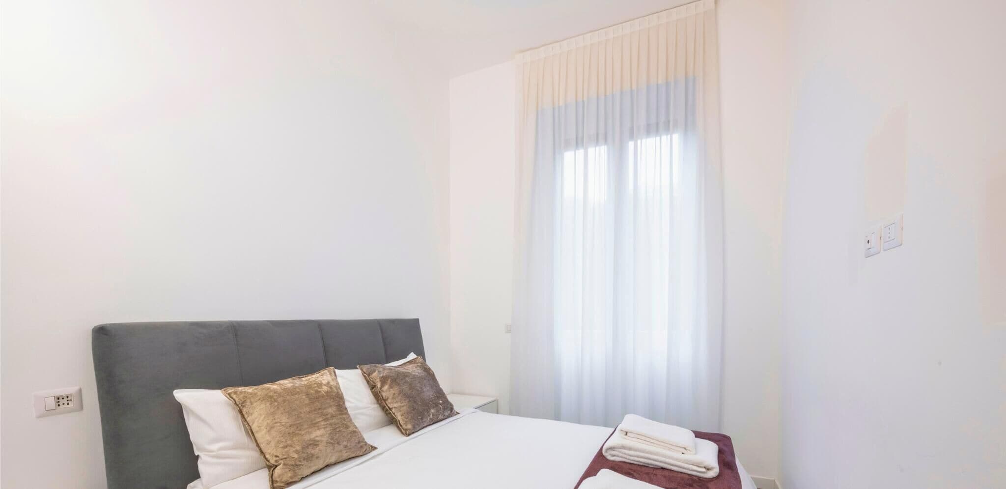 A bedroom with a bed, two pillows, a bedside table with a folded towel, and a window with sheer curtains.