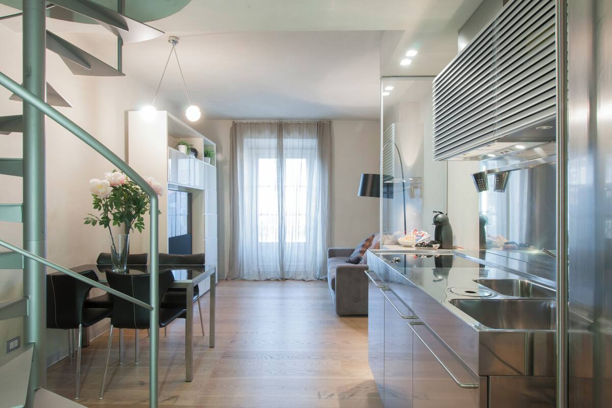 A modern kitchen with stainless steel appliances, a dining area with a glass table and chairs, and a staircase with metal railings.