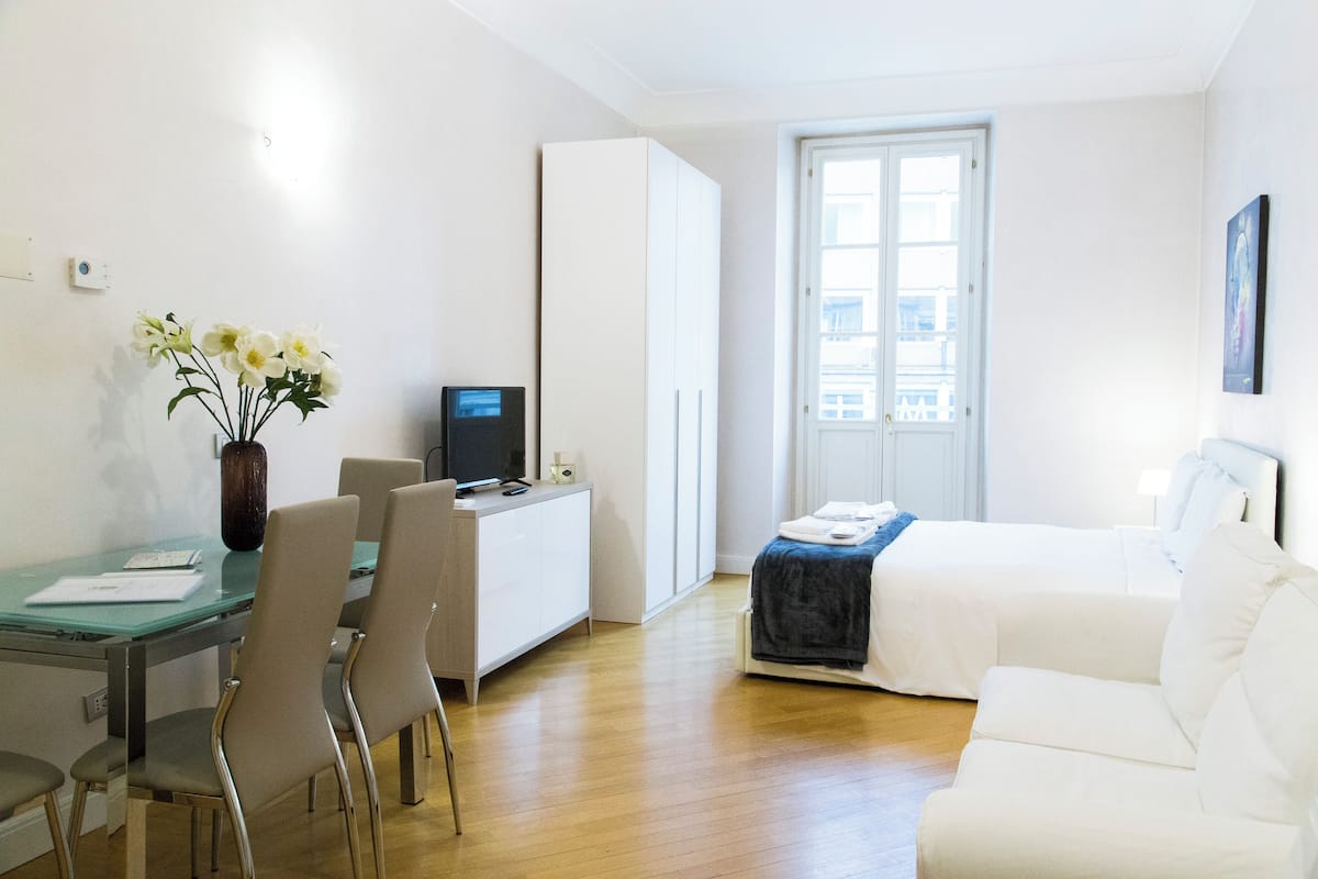 A hotel room with a bed, a desk with a computer, a chair, a small table with a vase of flowers, and a window with a view of buildings.