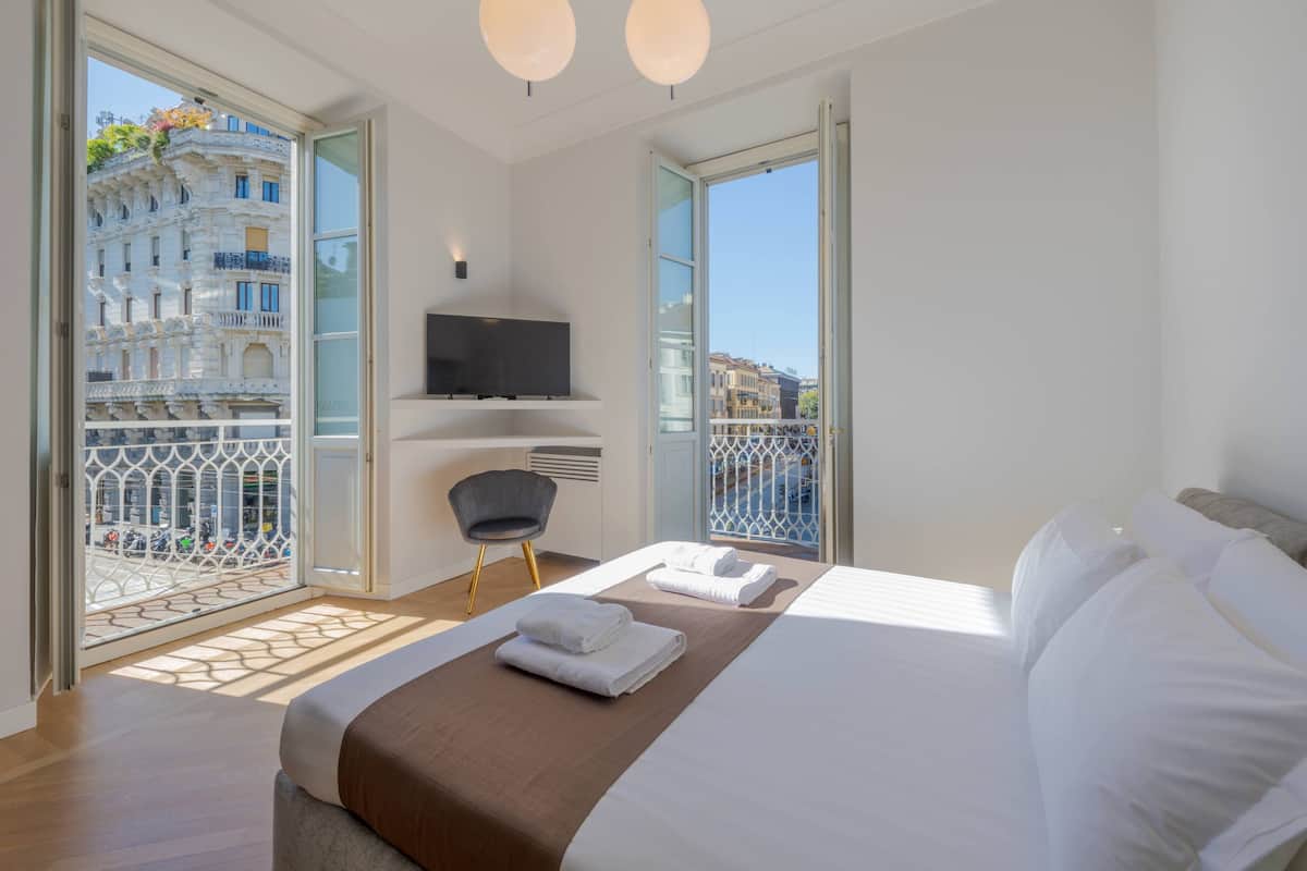 A modern hotel room with a large bed, a flat-screen TV, a balcony with a view of the city, and a chair.