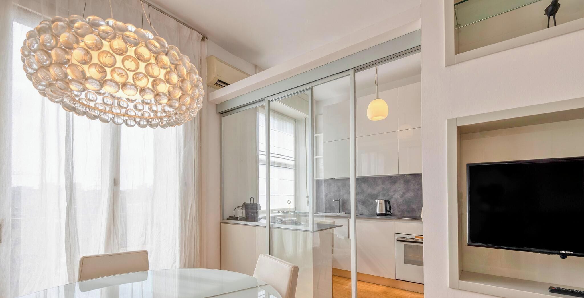 A modern kitchen with a glass partition, a dining area with a glass table, and a large flat-screen TV.