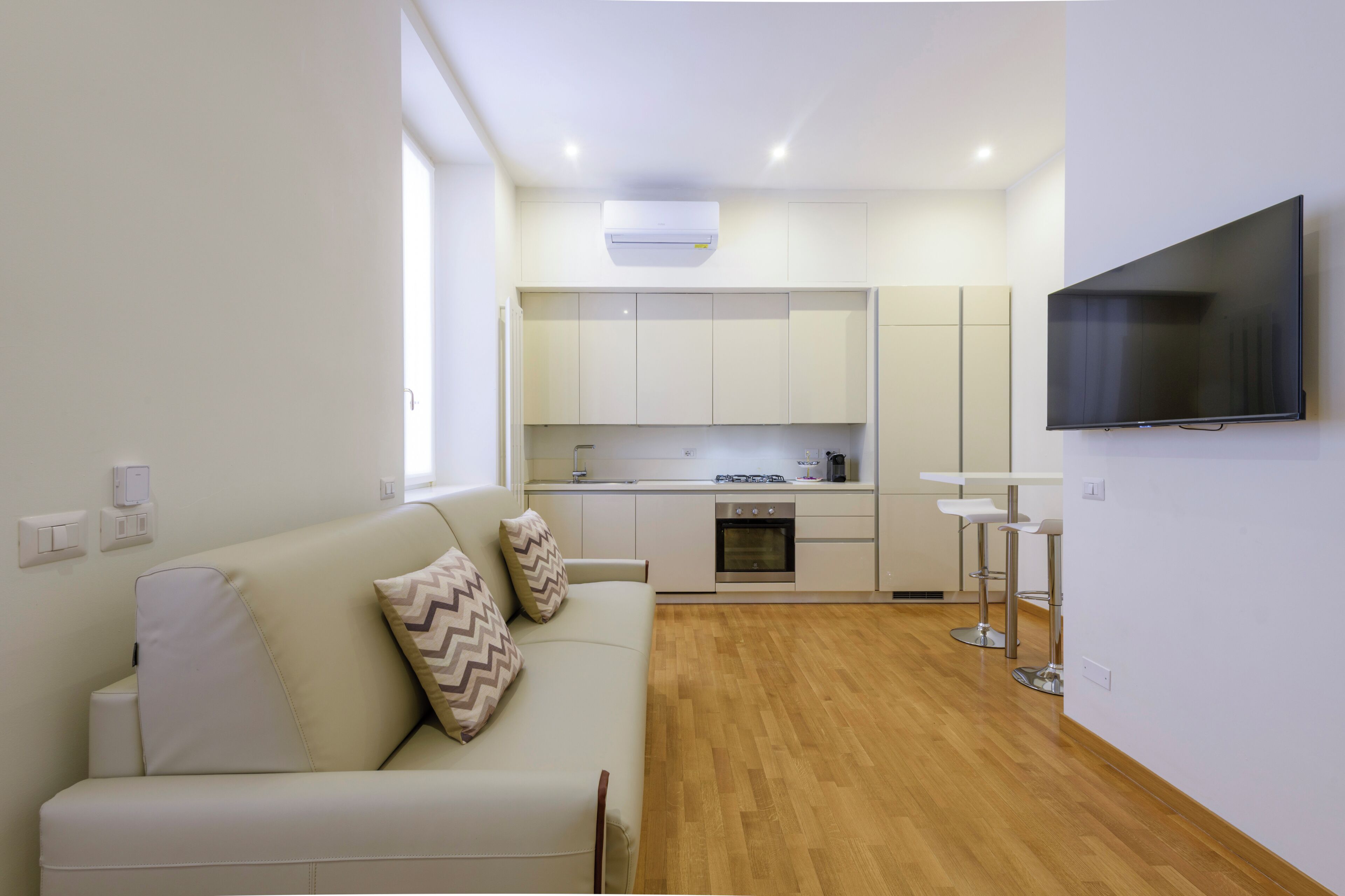 A modern living room with a sofa, a TV, a kitchen area with cabinets and a stove, and a dining area with stools.