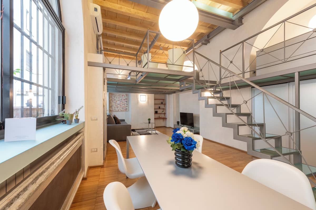 A modern interior with a dining area, white chairs, a table with a vase of flowers, a staircase with glass railings, and a large window.