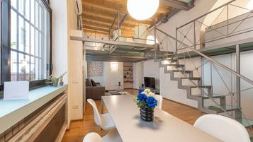 A modern interior with a dining area, white chairs, a table with a vase of flowers, a staircase with glass railings, and a large window.