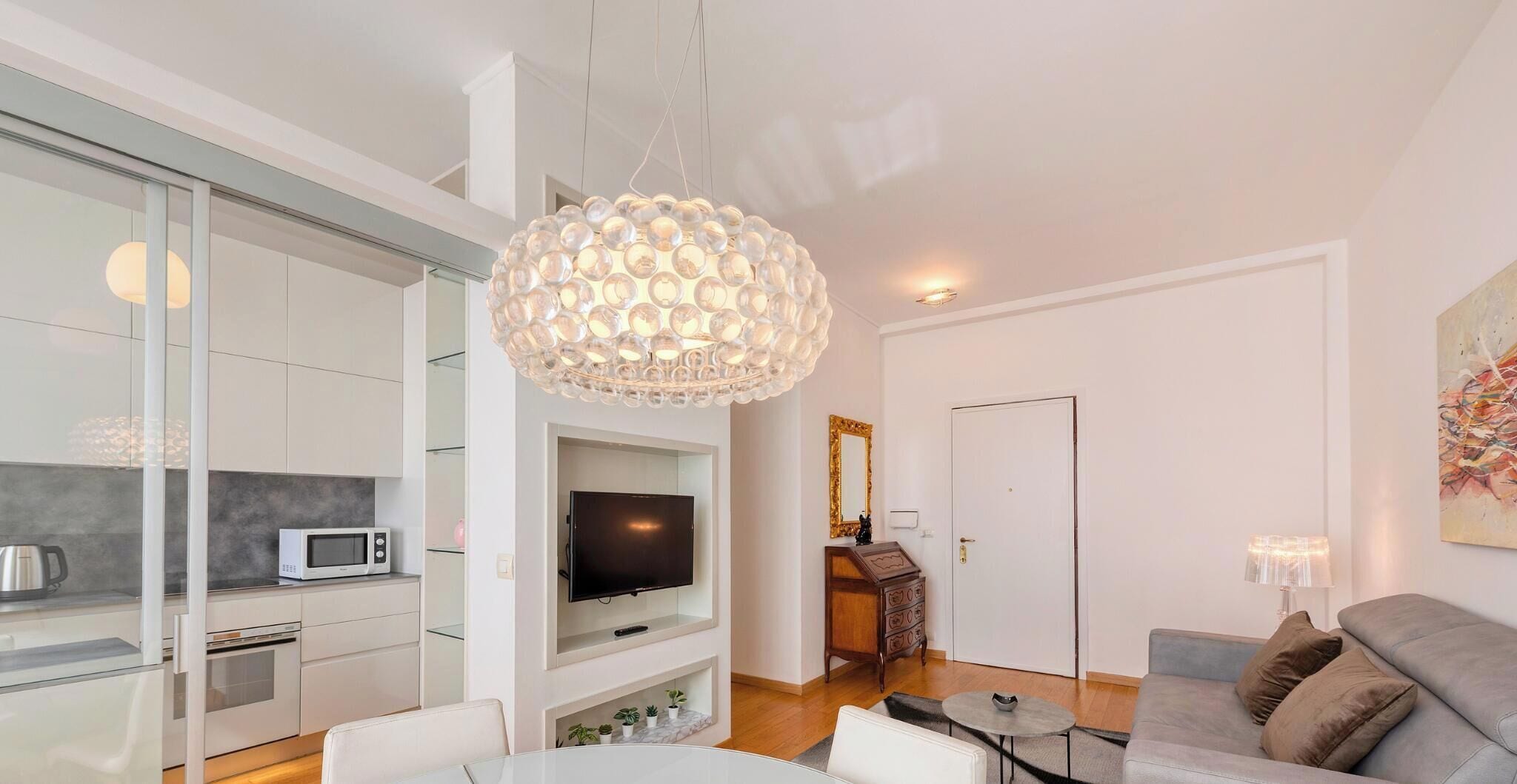 A modern living room with a grey sofa, a glass coffee table, and a television. There is a kitchen area with a microwave and a kettle. A unique chandelier hangs from the ceiling.