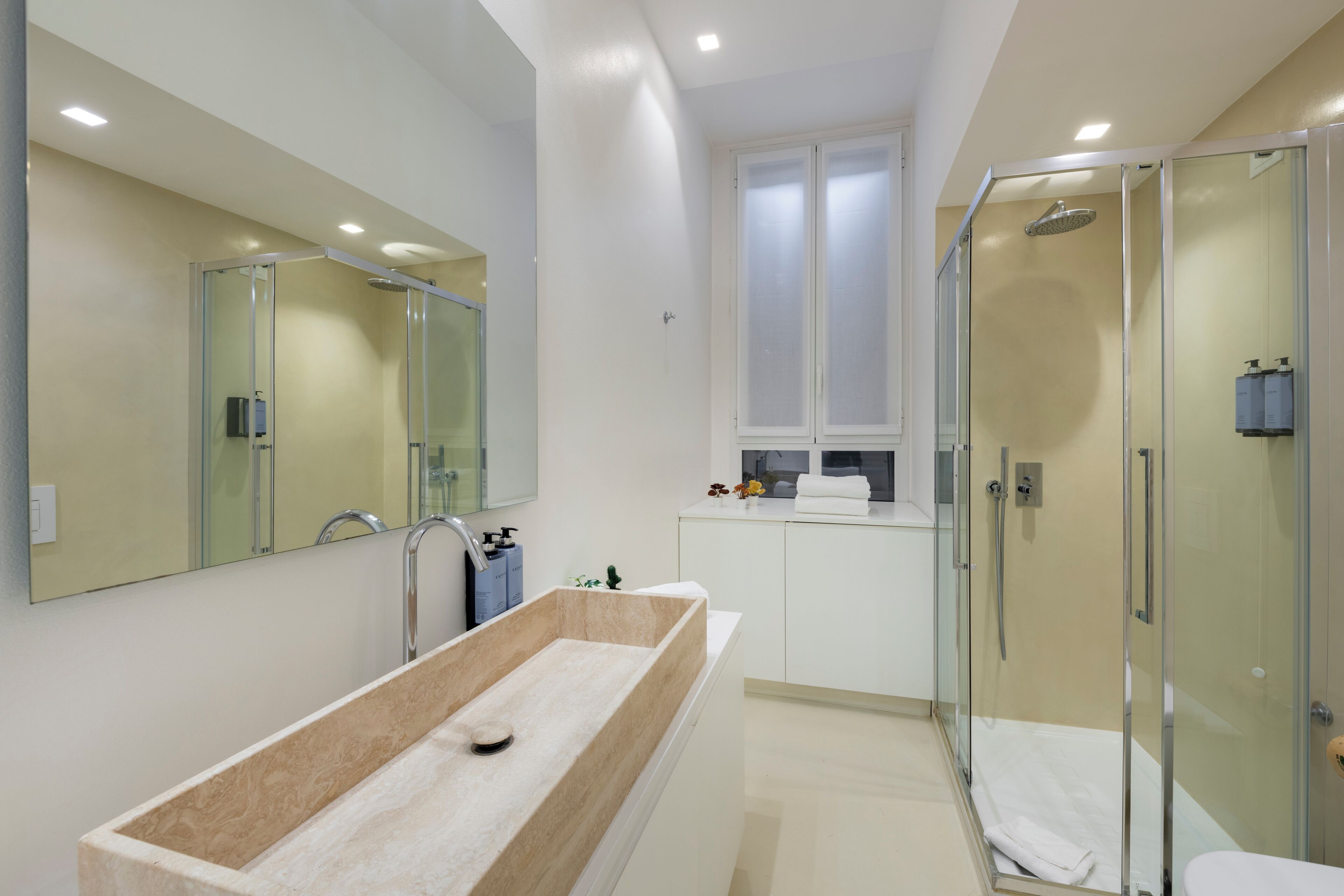 A modern bathroom with a large rectangular sink, a glass shower enclosure, and a window above the sink.