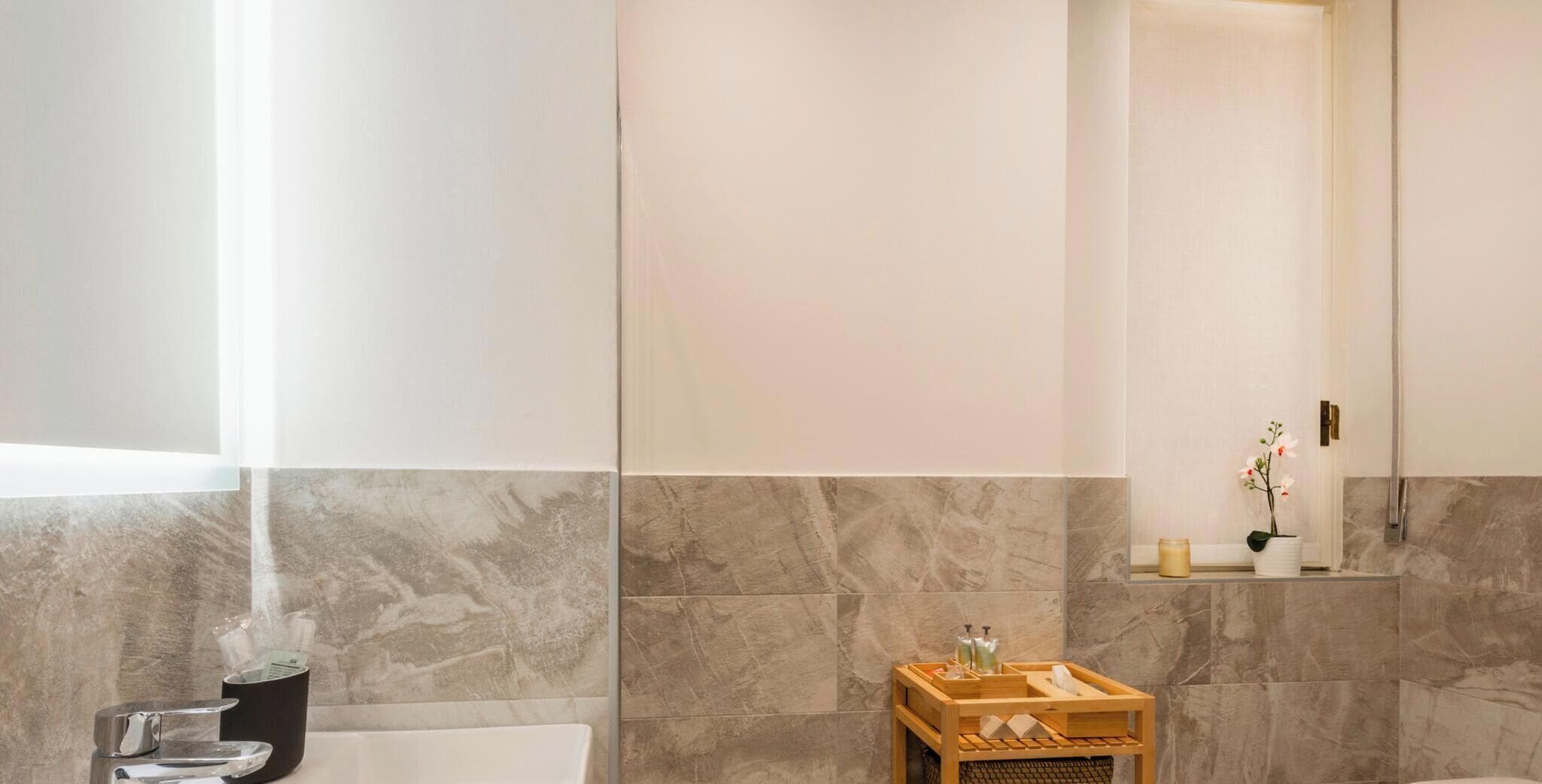 A modern bathroom with marble walls, a wooden vanity, and a glass shelf with toiletries.