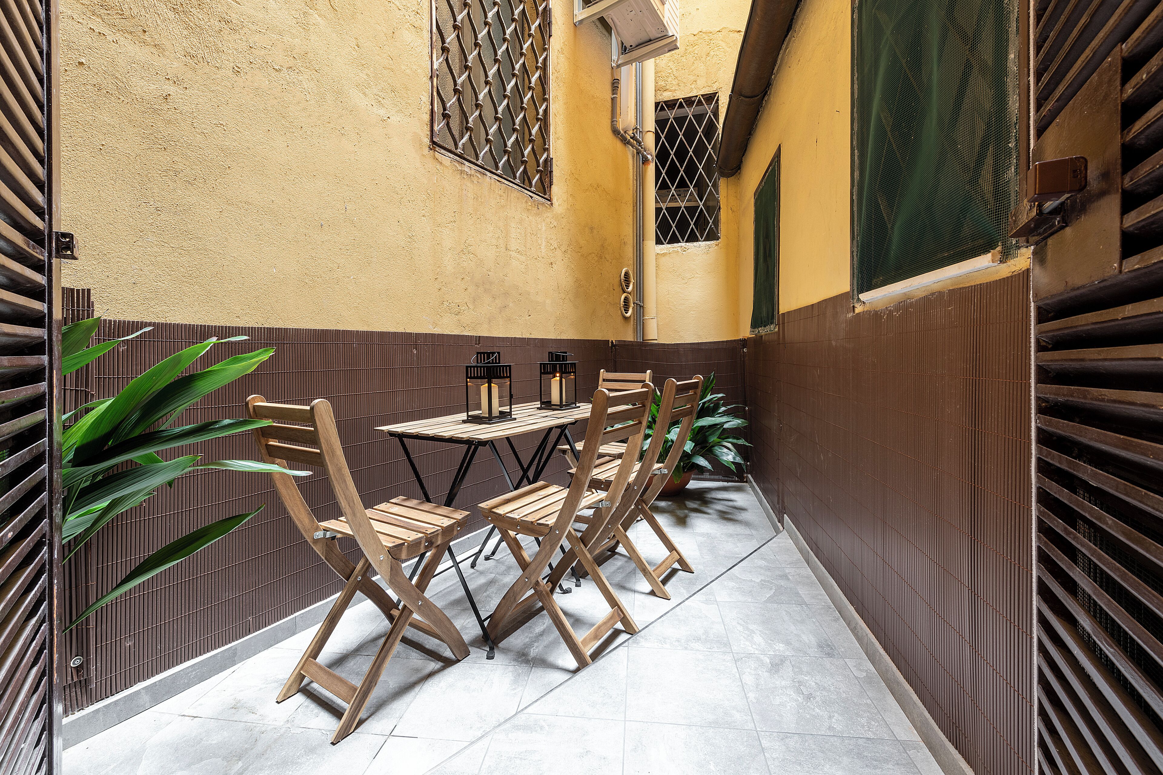 An outdoor patio with a table and chairs, a potted plant, and a wall with a window and ventilation system.