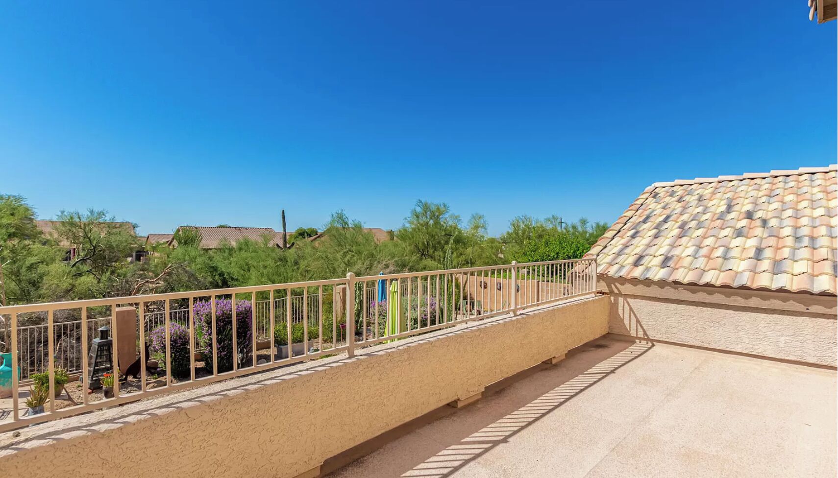 A balcony with a beige tiled floor, a metal railing, and a view of a residential area with houses and trees.