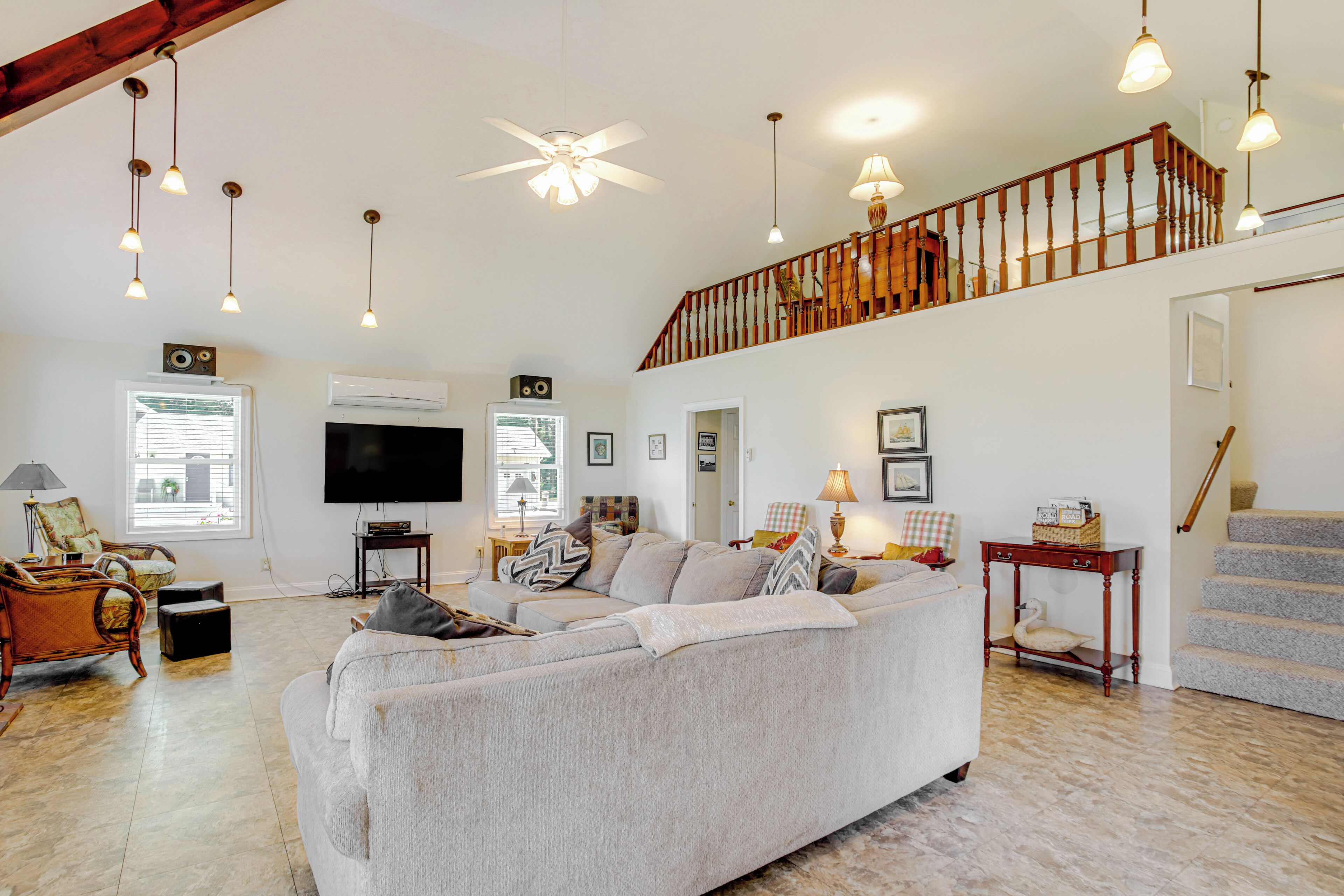 A spacious living room with a large grey sofa, wooden flooring, a ceiling fan, and a staircase leading to an upper level.