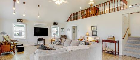 A spacious living room with a large grey sofa, wooden flooring, a ceiling fan, and a staircase leading to an upper level.