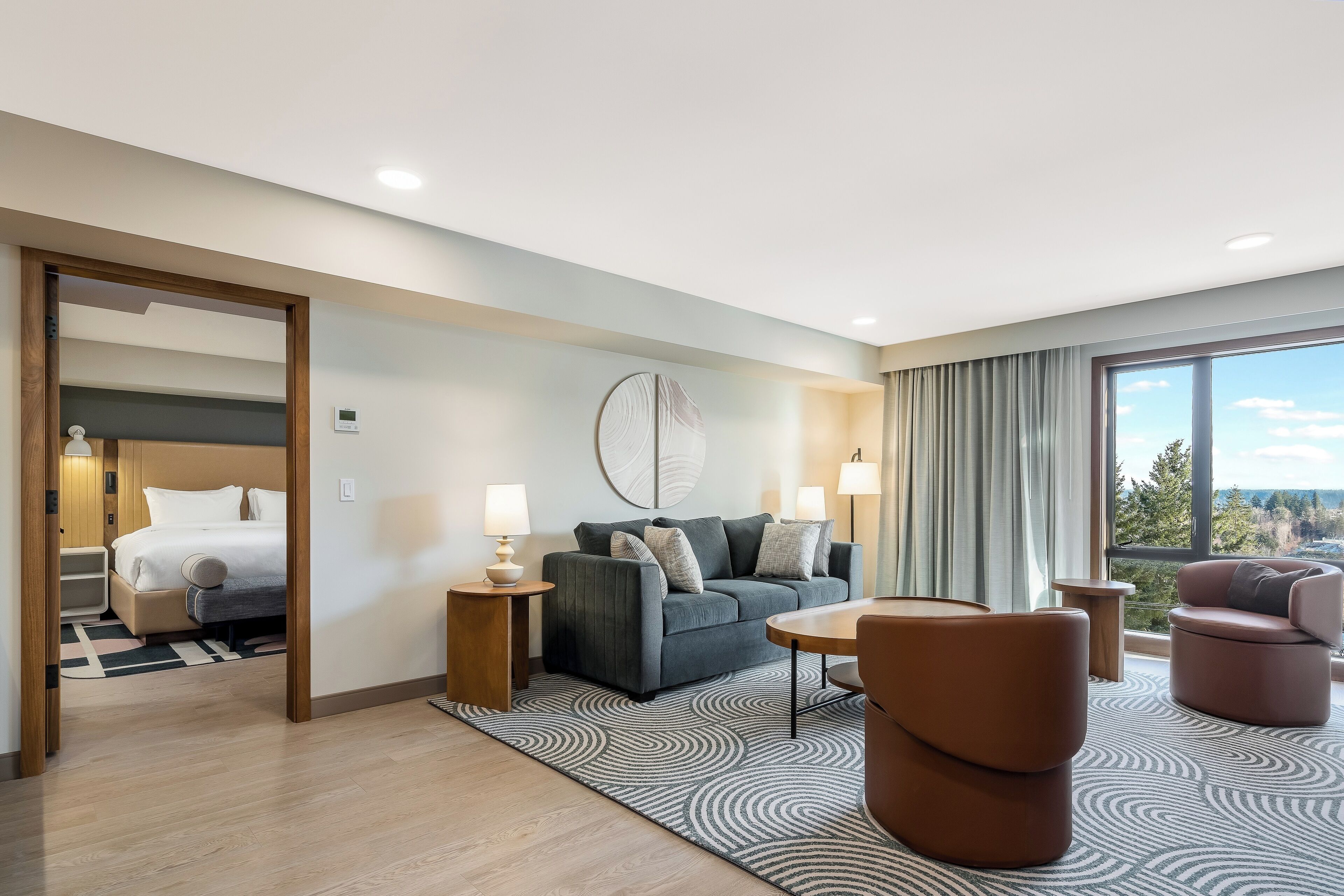 A modern hotel room with a bed, a grey sofa, two armchairs, a small table, and a large window offering a scenic view.