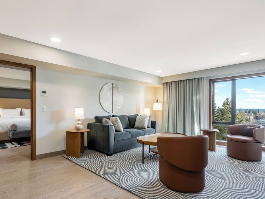 A modern hotel room with a bed, a grey sofa, two armchairs, a small table, and a large window offering a scenic view.