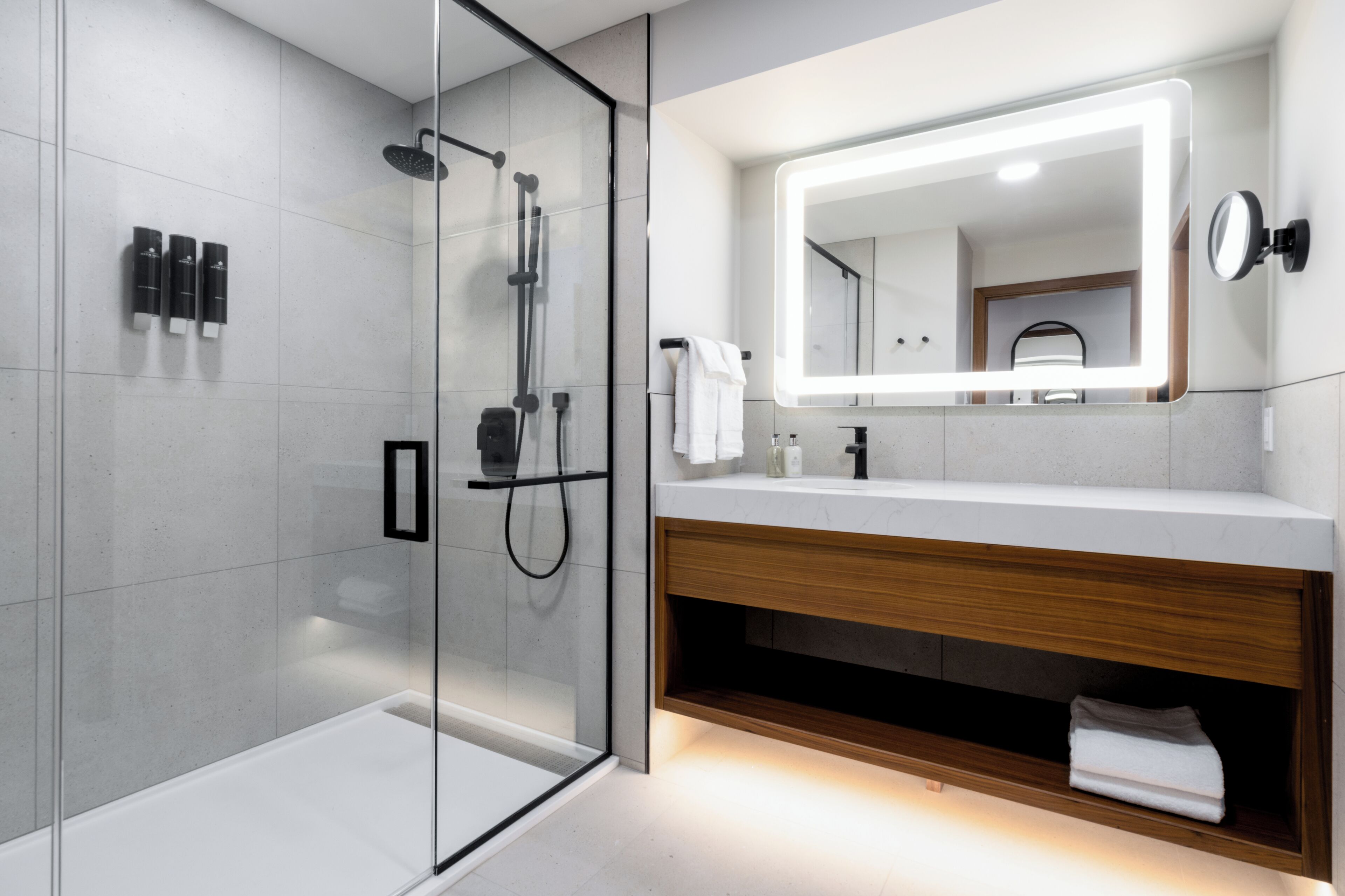 Un baño moderno con cabina de ducha acristalada, tocador con encimera de madera y un espejo grande con iluminación integrada.