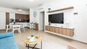 A modern living room with a TV, a dining area with a table and chairs, and a kitchen in the background. - Mary House by BarbarHouse (Talsano)