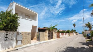 Una casa moderna de color blanco con un muro de piedra, una reja metálica y una vista despejada de la calle y el océano.