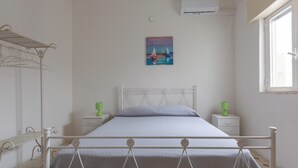 Un dormitorio con cama, dos mesitas de noche con lámparas, aire acondicionado de pared, una ventana y un cuadro en la pared.