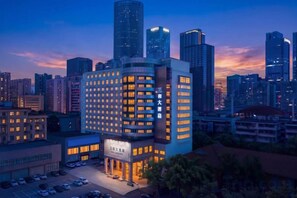 Un grand hôtel à plusieurs étages, avec un parking à l’avant et une vue sur les toits de la ville en arrière-plan.