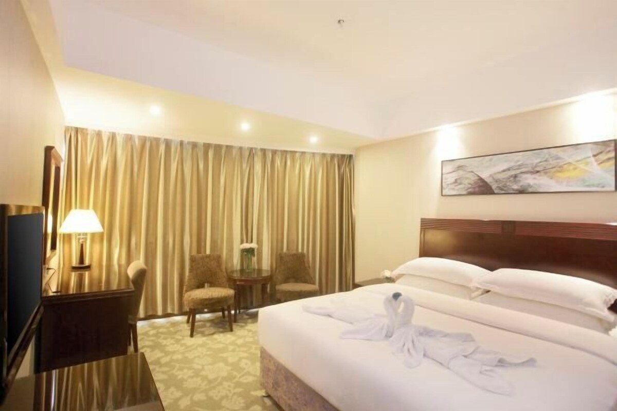 A hotel room with a large bed, a desk with a lamp, two chairs, and a painting on the wall.