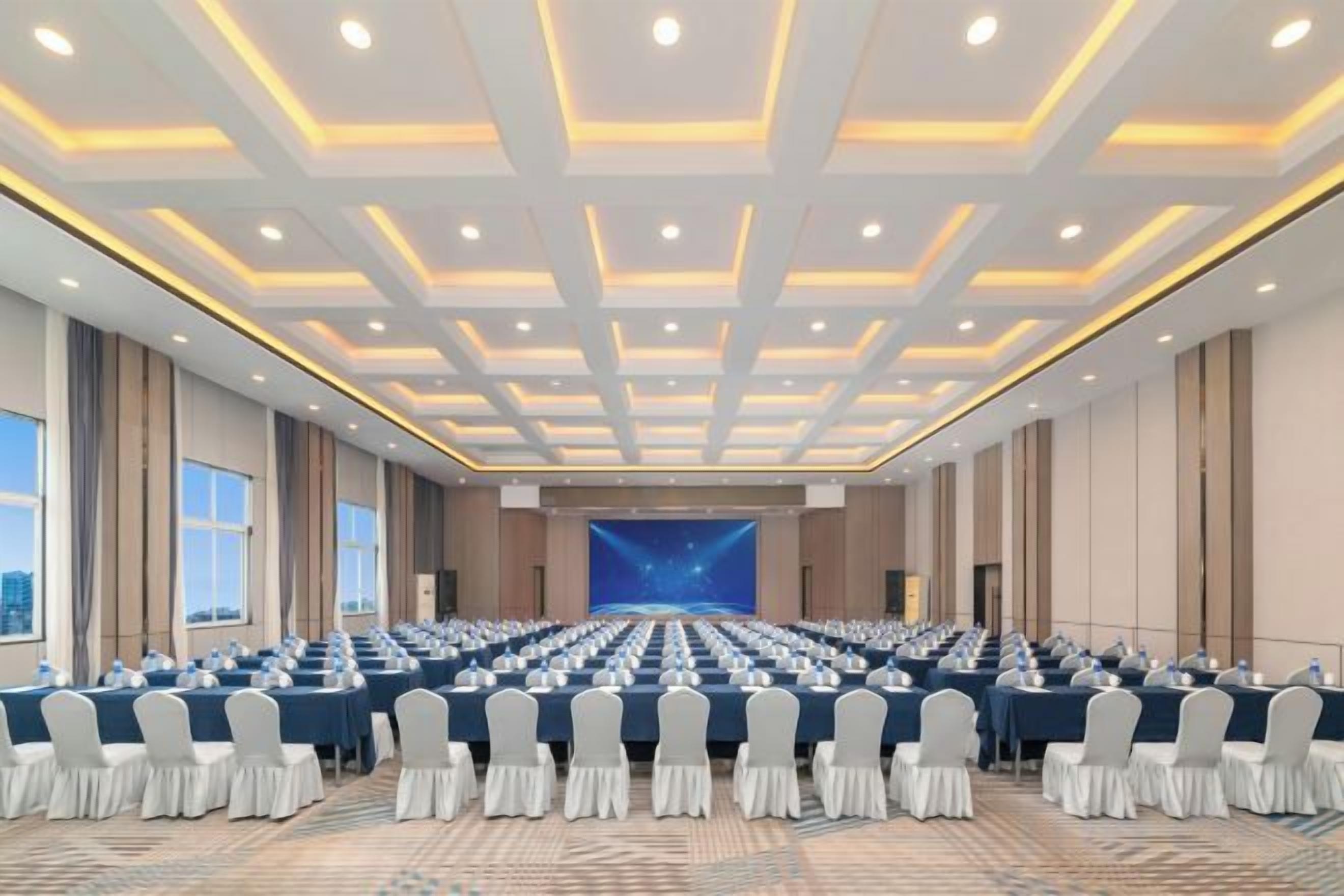 Un salón de conferencias con filas de mesas y sillas, una pantalla grande al frente y vistas de la ciudad a través de las ventanas.