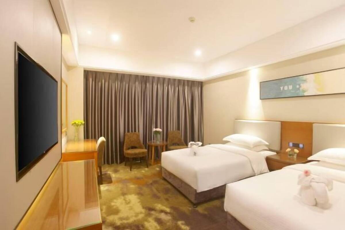 A hotel room with two beds, a desk with a chair, a TV, and a large window with curtains.